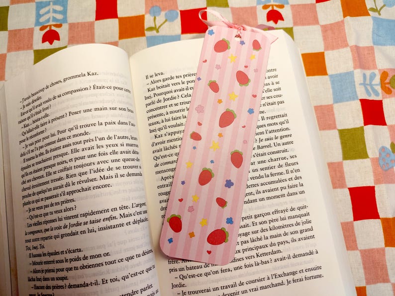 May include: A pink bookmark with a strawberry pattern, resting on an open book. The bookmark features red strawberries, yellow stars, and blue flowers. A pink ribbon is attached at the top.