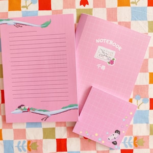 Op de afbeelding: Een roze notitieboek met een rasterpatroon en het woord "NOTEBOOK" gedrukt op de omslag. Het notitieboek heeft een sticker met een tekening van een draak en de tekst "千尋".