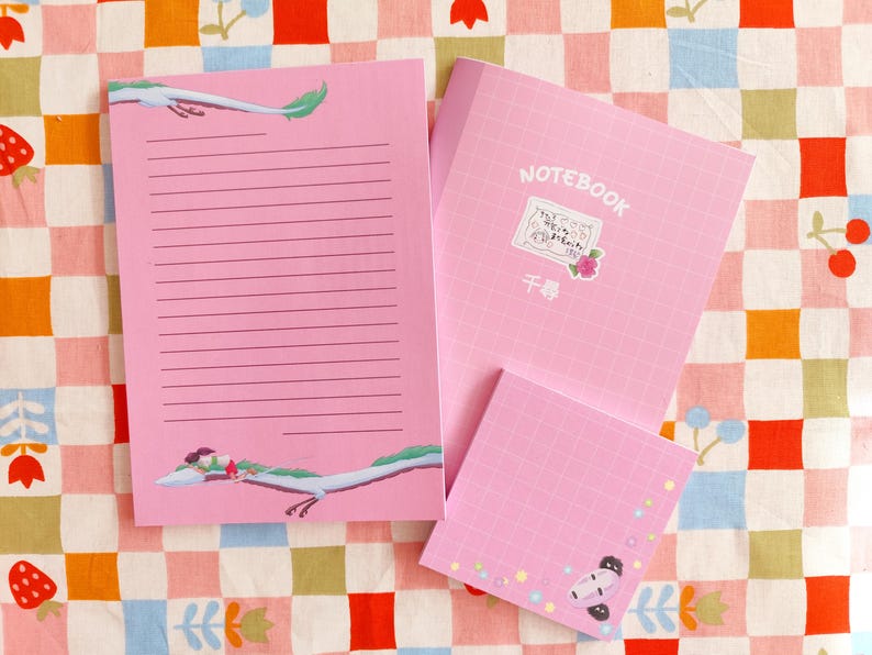 Op de afbeelding: Een roze notitieboek met een rasterpatroon en het woord "NOTEBOOK" gedrukt op de omslag. Het notitieboek heeft een sticker met een tekening van een draak en de tekst "千尋".