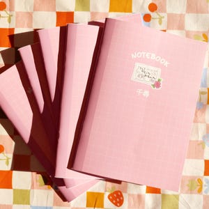 Op de afbeelding: Een stapel roze notitieboekjes met een rasterpatroon op de kaft. Het bovenste notitieboekje heeft het woord "NOTEBOOK" op de kaft gedrukt, samen met een kleine sticker met de tekst "千尋".