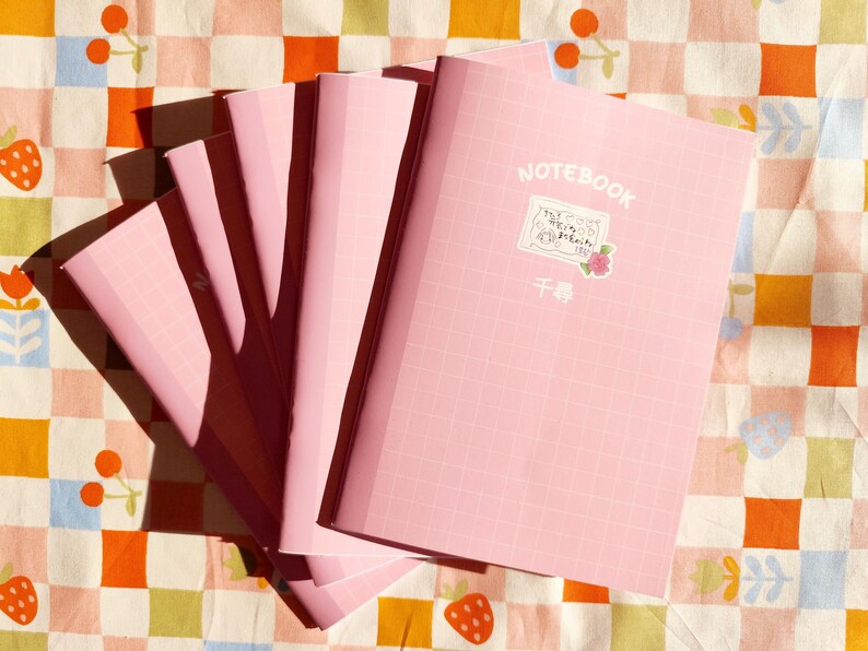 Op de afbeelding: Een stapel roze notitieboekjes met een rasterpatroon op de kaft. Het bovenste notitieboekje heeft het woord "NOTEBOOK" op de kaft gedrukt, samen met een kleine sticker met de tekst "千尋".