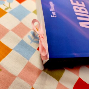 May include: A blue book with the title "AUBE" and the author's name, Eve Hauguin, is shown with a pink ribbon bookmark. The book rests on a colorful patterned fabric with a strawberry and floral design.