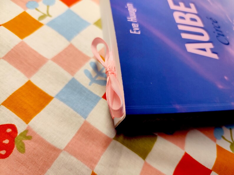 May include: A blue book with the title "AUBE" and the author's name, Eve Hauguin, is shown with a pink ribbon bookmark. The book rests on a colorful patterned fabric with a strawberry and floral design.