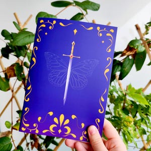 May include: A greeting card with a dark blue background and a gold decorative border. A silver sword is centered over a butterfly outline. The sword has a gold hilt with a red gem. The card is held in front of a green leafy background.