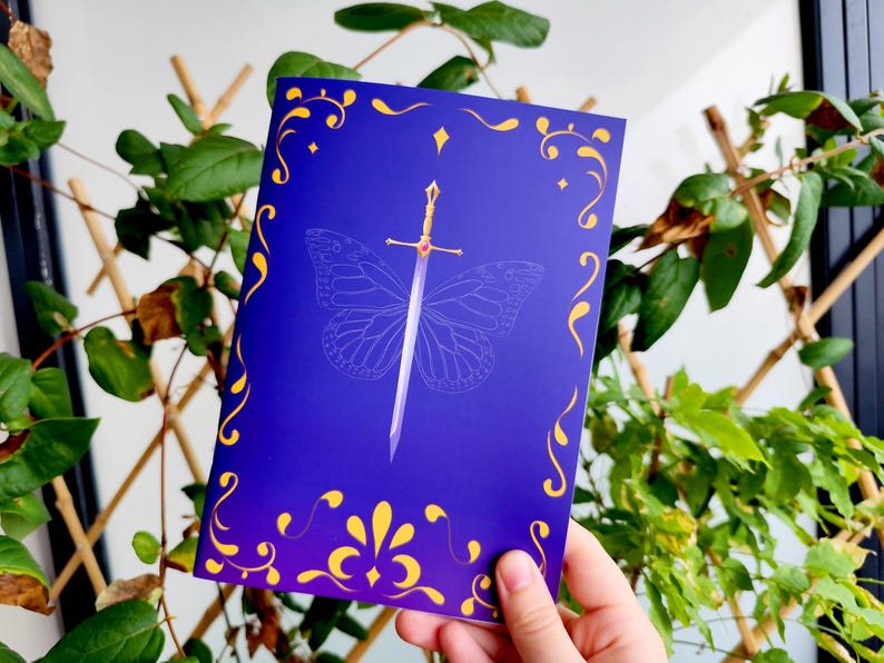 May include: A greeting card with a dark blue background and a gold decorative border. A silver sword is centered over a butterfly outline. The sword has a gold hilt with a red gem. The card is held in front of a green leafy background.