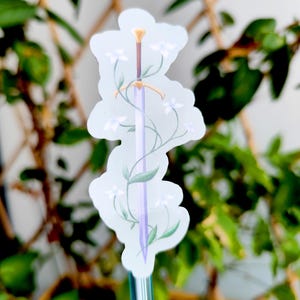 May include: A sticker featuring a sword design with a light purple blade, a brown handle, and a gold hilt. The sword is adorned with white flowers and green leaves. The sticker has a white background and is surrounded by green foliage.