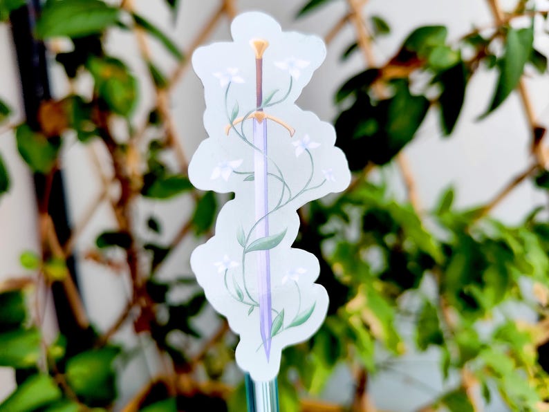 May include: A sticker featuring a sword design with a light purple blade, a brown handle, and a gold hilt. The sword is adorned with white flowers and green leaves. The sticker has a white background and is surrounded by green foliage.