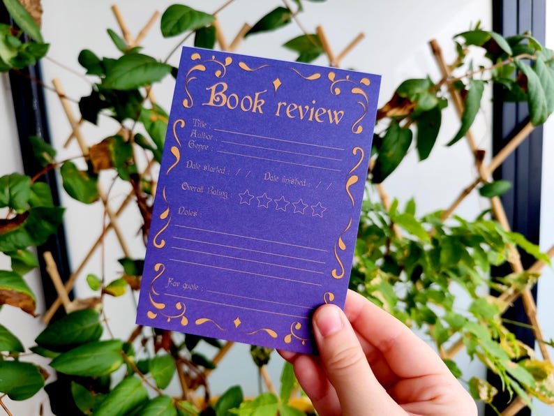 May include: A purple book review card with gold decorative accents and text. The card includes fields for title, author, genre, dates, rating, and notes. The card is held in front of a background of green leaves.