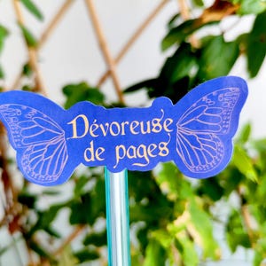 May include: A blue butterfly-shaped bookmark with the French text "D&eacute;voreuse de pages" in gold lettering. The butterfly wings have a delicate, light purple outline. The bookmark is attached to a clear, green-tinted stick, set against a backdrop of green foliage.