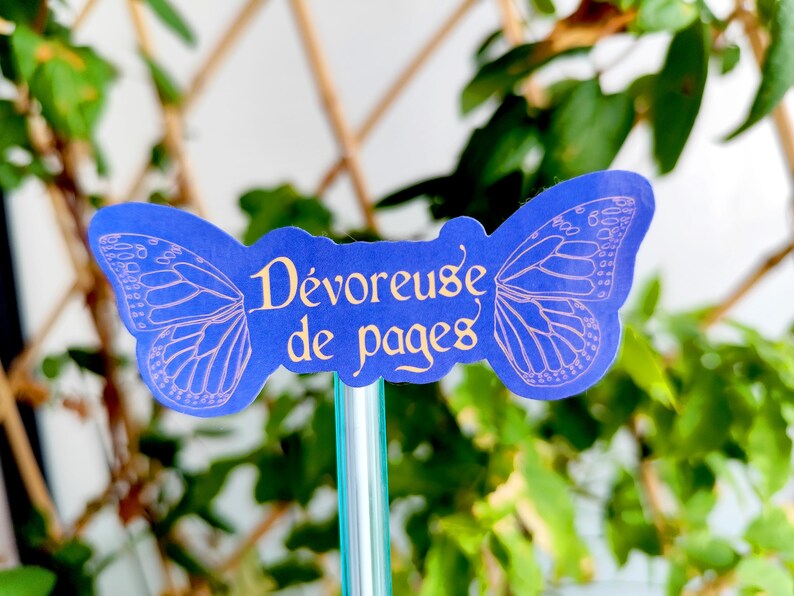 May include: A blue butterfly-shaped bookmark with the French text "D&eacute;voreuse de pages" in gold lettering. The butterfly wings have a delicate, light purple outline. The bookmark is attached to a clear, green-tinted stick, set against a backdrop of green foliage.