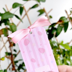 May include: A pink gift tag with a striped pattern and egg-shaped designs, tied with a matching pink ribbon. The tag has a small hole at the top for the ribbon. The background features green foliage.