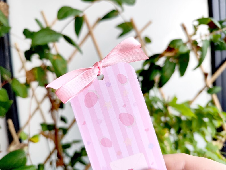 May include: A pink gift tag with a striped pattern and egg-shaped designs, tied with a matching pink ribbon. The tag has a small hole at the top for the ribbon. The background features green foliage.