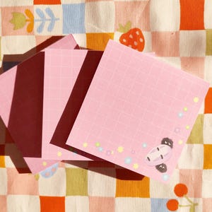 Op de afbeelding: Drie roze plakbriefjes met een rasterpatroon en een cartoonfiguur op het grootste briefje. De briefjes liggen op een geruite achtergrond met roze, oranje en witte vierkanten.
