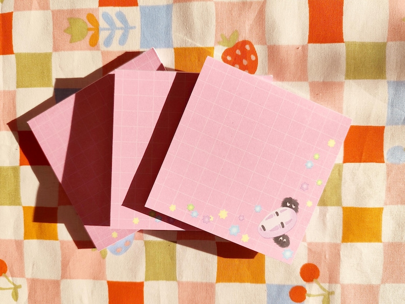 Op de afbeelding: Drie roze plakbriefjes met een rasterpatroon en een cartoonfiguur op het grootste briefje. De briefjes liggen op een geruite achtergrond met roze, oranje en witte vierkanten.