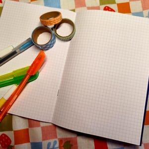 May include: An open notebook with grid paper, surrounded by colorful pens and decorative tape rolls. The pens are in shades of blue, green, orange, and yellow. The tape rolls feature patterns like gingham and polka dots.