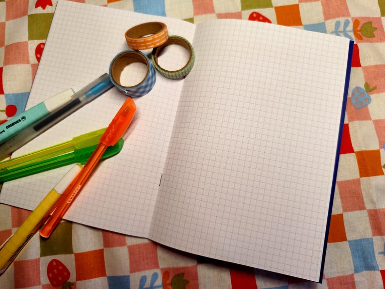 May include: An open notebook with grid paper, surrounded by colorful pens and decorative tape rolls. The pens are in shades of blue, green, orange, and yellow. The tape rolls feature patterns like gingham and polka dots.