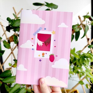 Peut inclure: Carnet rose à rayures verticales, avec une image de style polaroid de deux mains se tenant. Le carnet est décoré d'illustrations de nuages et d'étoiles, d'une fraise et d'une fleur.