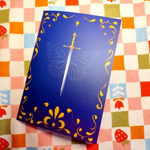 May include: A blue book cover with a gold sword design and a butterfly outline. The cover has gold decorative flourishes and is set against a colorful checkered background with strawberries and flowers.