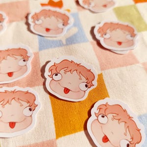 Puede incluir: Un conjunto de ocho pegatinas de personajes de dibujos animados con fondo blanco y detalles en rosa y rojo. Los personajes tienen ojos grandes y sacan la lengua. Las pegatinas est&aacute;n sobre un fondo a cuadros.