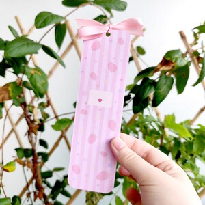 May include: A pink bookmark with vertical stripes and scattered pink ovals. A small envelope graphic with a heart is centered on the bookmark. A pink ribbon is attached at the top. The bookmark is held in front of a background of green plants.