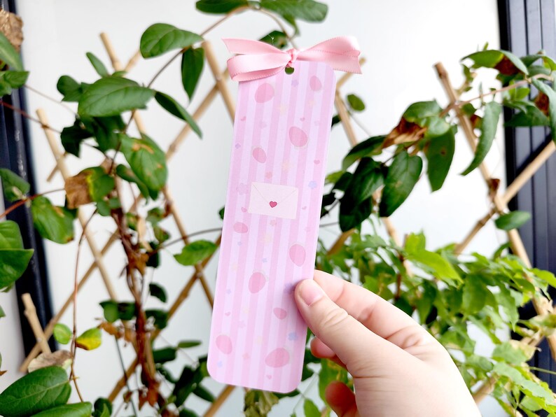 May include: A pink bookmark with vertical stripes and scattered pink ovals. A small envelope graphic with a heart is centered on the bookmark. A pink ribbon is attached at the top. The bookmark is held in front of a background of green plants.