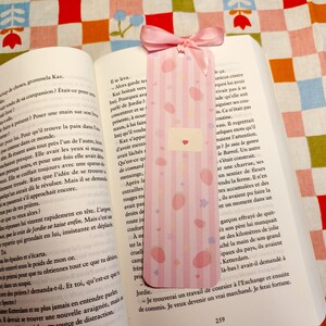 May include: A pink bookmark with vertical stripes, pink polka dots, and a small envelope design with a heart, resting in an open book. A pink ribbon is attached to the top. The book is on a colorful patterned surface.