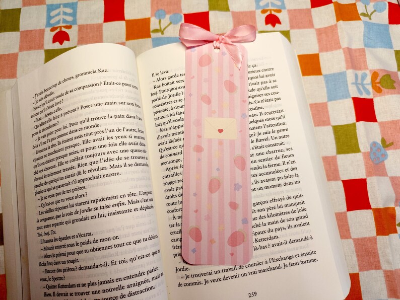 May include: A pink bookmark with vertical stripes, pink polka dots, and a small envelope design with a heart, resting in an open book. A pink ribbon is attached to the top. The book is on a colorful patterned surface.