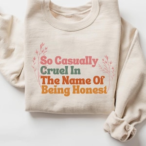May include: A beige sweatshirt with the text "So Casually Cruel In The Name Of Being Honest" printed on the front in colourful letters. The text is surrounded by small pink floral designs.