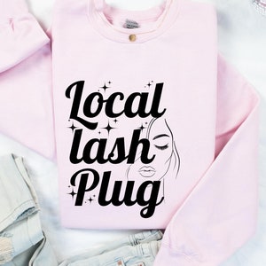 May include: A light pink sweatshirt with the words "Local Lash Plug" in black script, along with a line drawing of a woman's face with closed eyes and full lips. The sweatshirt is folded, with a pair of light-wash jeans visible.