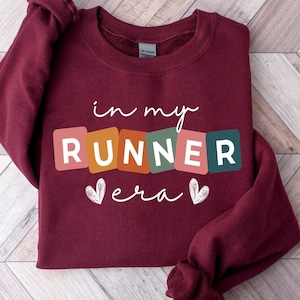 May include: Burgundy sweatshirt with the text "in my runner era" in colorful block letters with white hearts on either side.
