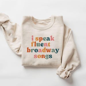 Könnte beinhalten: Cremefarbenes Sweatshirt mit dem Text "i speak fluent broadway songs" in bunten Buchstaben im Retro-Stil. Die langen Ärmel sind hochgekrempelt. Ein lässiges, bequemes Kleidungsstück für Musical-Fans.