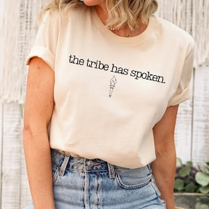 May include: Cream-colored t-shirt with the phrase "the tribe has spoken" printed in black, along with a torch graphic. The shirt has rolled-up sleeves and is paired with blue denim jeans. The overall style is casual and comfortable.