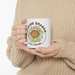 Puede incluir: Taza de cerámica blanca con el texto "Jasmine Dragon Tea House" en un diseño circular, con una ilustración de tetera. El diseño incluye hojas verdes y flores blancas, con las palabras "Ba Sing Se" en el lateral.