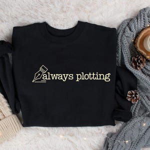 May include: Black crew neck sweatshirt with the text "always plotting" in a cream-colored font. The design includes a pen nib illustration. The sweatshirt is displayed with a knitted beanie, a gray cable-knit blanket, pine cones, and a candle.