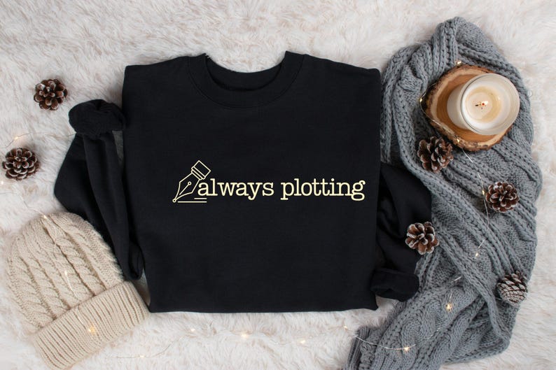 May include: Black crew neck sweatshirt with the text "always plotting" in a cream-colored font. The design includes a pen nib illustration. The sweatshirt is displayed with a knitted beanie, a gray cable-knit blanket, pine cones, and a candle.