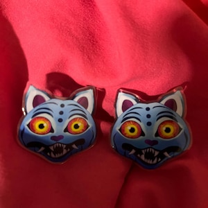 Peut inclure: Deux épingles en émail en forme de chat. Chaque épingle présente un visage de chat bleu avec de grands yeux jaunes, des dents pointues et un contour argenté. Les épingles sont posées sur un fond rouge.