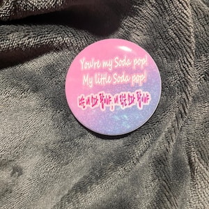 May include: A round button with a pink and purple gradient background. The button has the text "You're my Soda pop! My Little Soda pop!" in white. Below the English text is text in a different language.