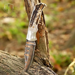 May include: A handcrafted knife with a patterned blade and a handle made of bone and metal. The knife is displayed against a natural wood background. The text "Rayah Knives" is visible.