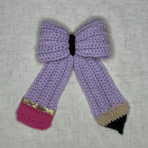 May include: A handmade crochet bow in the shape of a pencil. The bow is lavender with a black center. The pencil tips are pink and tan with gold glitter accents and a black tip.