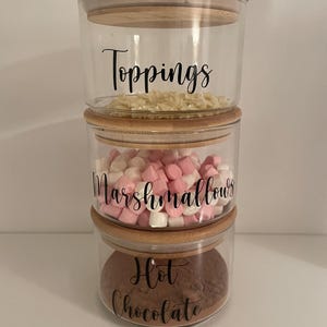 May include: Three glass jars with bamboo lids, stacked on top of each other. The jars are labelled "Toppings", "Marshmallows", and "Hot Chocolate".