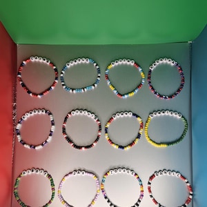 May include: A collection of colorful beaded bracelets with white letter beads spelling out various names. Each bracelet features a unique color scheme, including red, blue, yellow, and green, with a mix of other colors.