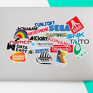 May include: A laptop adorned with a collection of stickers displaying logos of retro video game companies, including Atari, Sega, Nintendo, and Capcom. The stickers are arranged in a collage, with various shapes and sizes. The laptop is on a white surface.