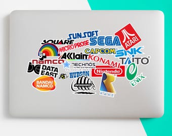 May include: A laptop covered in colorful stickers featuring logos of classic video game companies like Atari, Sega, Nintendo, and Capcom. The stickers are arranged in a collage, showcasing a variety of shapes and sizes. The laptop is on a white surface.