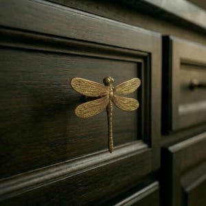 May include: A bronze dragonfly cabinet pull is fixed to a dark wood cabinet door. The dragonfly has detailed wings and a long, slender body. The hardware adds a decorative touch to the cabinet.