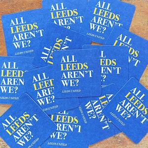 May include: A pile of blue square coasters, each printed with the text "ALL LEEDS AREN'T WE?" in yellow and white. The words "LEEDS UNITED" are printed at the bottom. The coasters are arranged on a wooden surface.