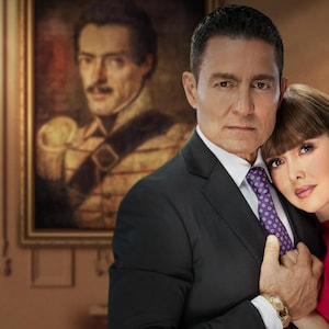 K&ouml;nnte beinhalten: Ein Paar posiert f&uuml;r ein Portr&auml;t in einem formellen Ambiente. Der Mann tr&auml;gt einen dunklen Anzug mit einer violetten Krawatte. Die Frau tr&auml;gt ein rotes Kleid. Ein Portr&auml;t eines Mannes in Milit&auml;runiform h&auml;ngt an der Wand hinter ihnen.