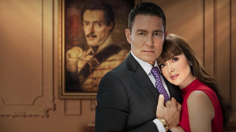 K&ouml;nnte beinhalten: Ein Paar posiert f&uuml;r ein Portr&auml;t in einem formellen Ambiente. Der Mann tr&auml;gt einen dunklen Anzug mit einer violetten Krawatte. Die Frau tr&auml;gt ein rotes Kleid. Ein Portr&auml;t eines Mannes in Milit&auml;runiform h&auml;ngt an der Wand hinter ihnen.