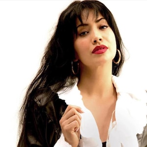 May include: A woman with long dark hair and a white blouse with a ruffled collar. She is wearing a brown leather jacket and has large gold hoop earrings. She is looking at the camera with a serious expression.