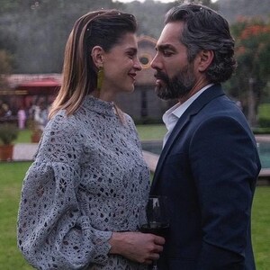 May include: A couple standing outdoors in front of a pool. The woman is wearing a light blue crocheted top and the man is wearing a dark blue suit jacket over a white shirt.