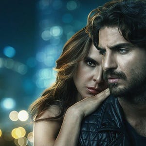 May include: A man and a woman stand close together in a dimly lit urban setting. The man is wearing a black leather jacket and has a beard. The woman has long brown hair and is wearing a black top. The background is blurred and features out-of-focus lights.